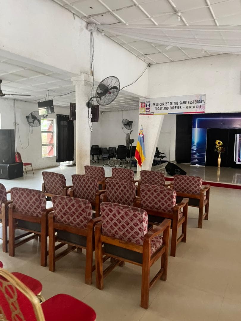 Inside the current auditorium — wooden chairs, standing fans and a modest stage.